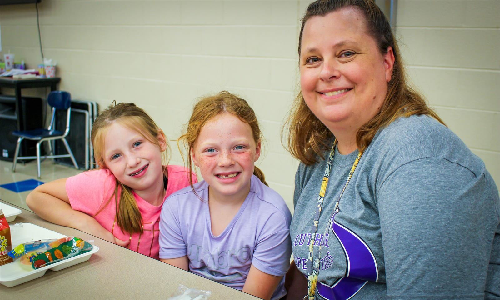 A teacher smiles while sitting beside to young girls at a lunch table